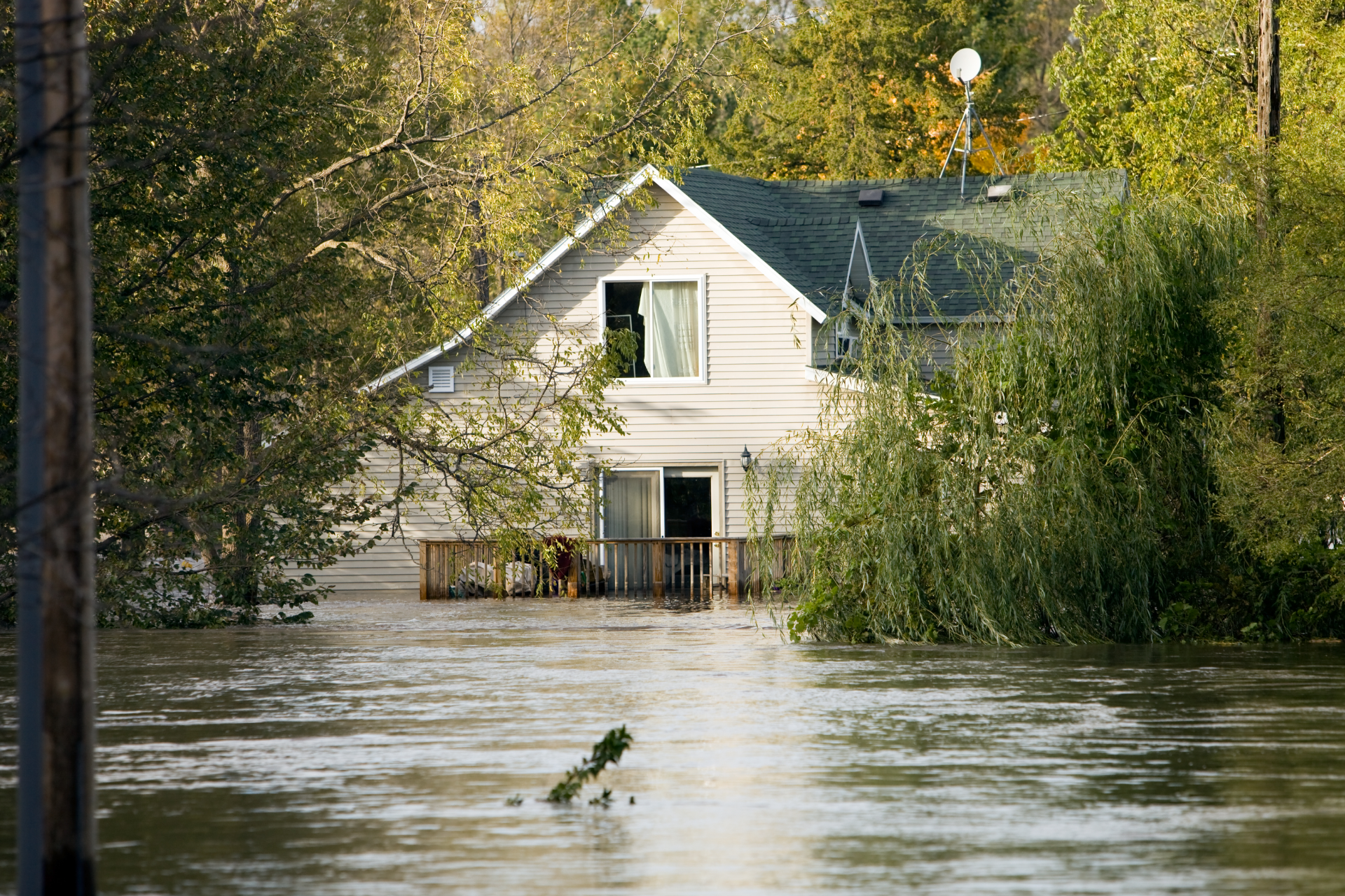 A single house surrounded by rising floodwaters, with its lower level partially submerged and trees in the background.