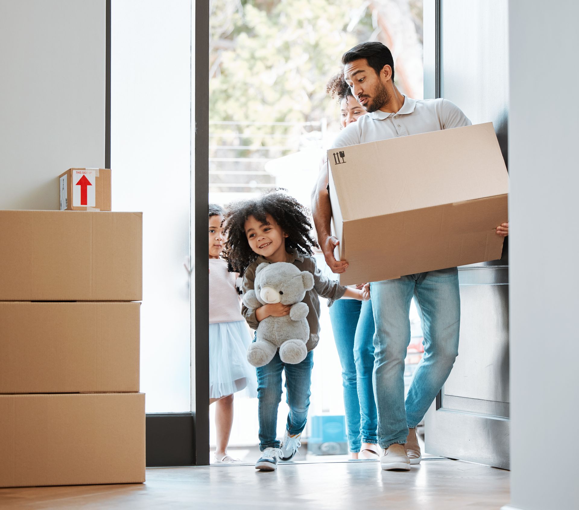 A family enters a new home carrying cardboard moving boxes and a stuffed animal.