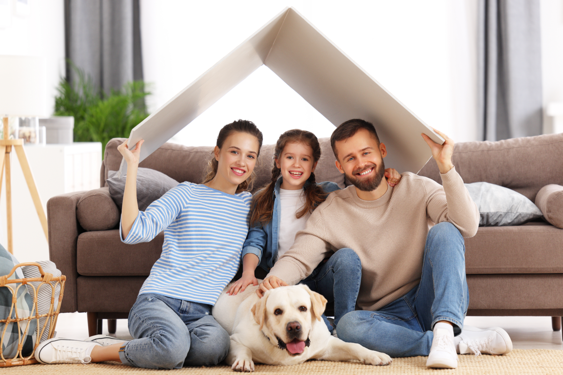 A family and a dog sit together on a living room floor under a symbolic roof made from two pieces of white board.