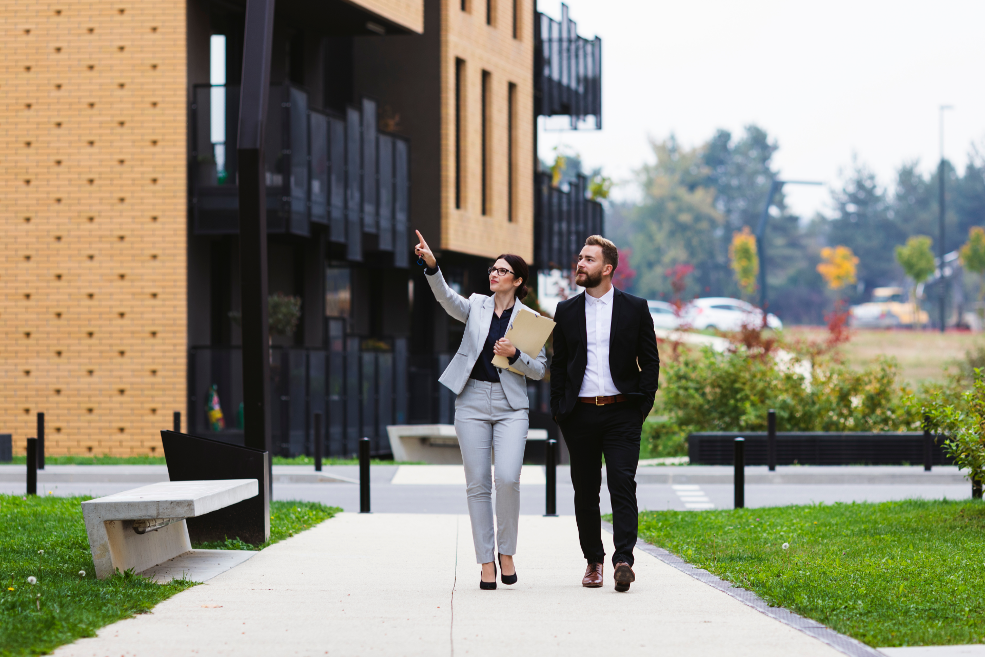 Two professionals in business attire walk on a path outside a modern building, one pointing toward the structure.