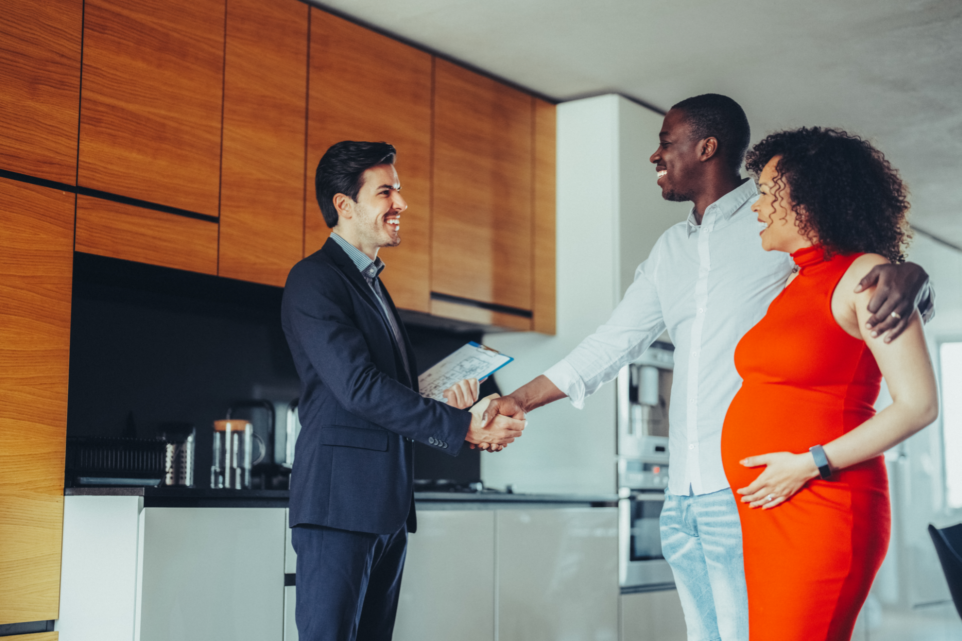 A professional in a suit shakes hands with a man, while a pregnant woman in a red dress smiles, standing in a kitchen.