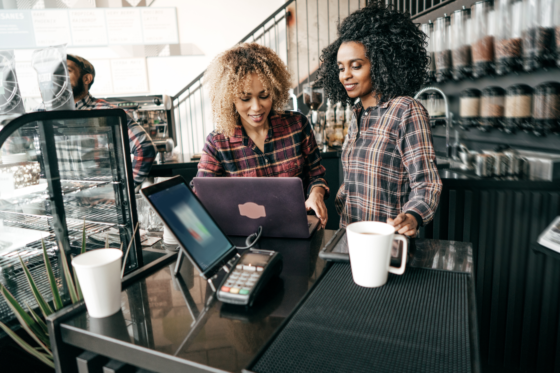 Two people in plaid shirts collaborate at a coffee shop counter with a laptop, payment terminal, and coffee mugs.