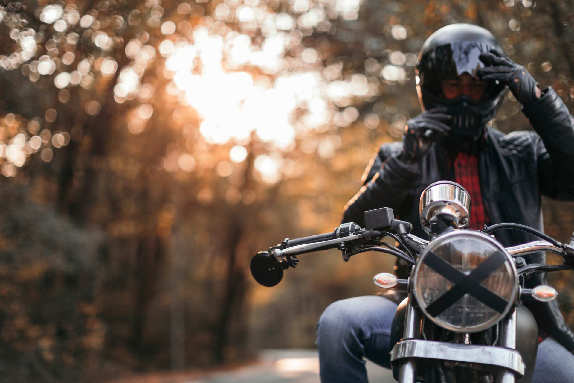 A motorcyclist in a leather jacket adjusts a black helmet while sitting on a bike on a road lined with sunlit trees.