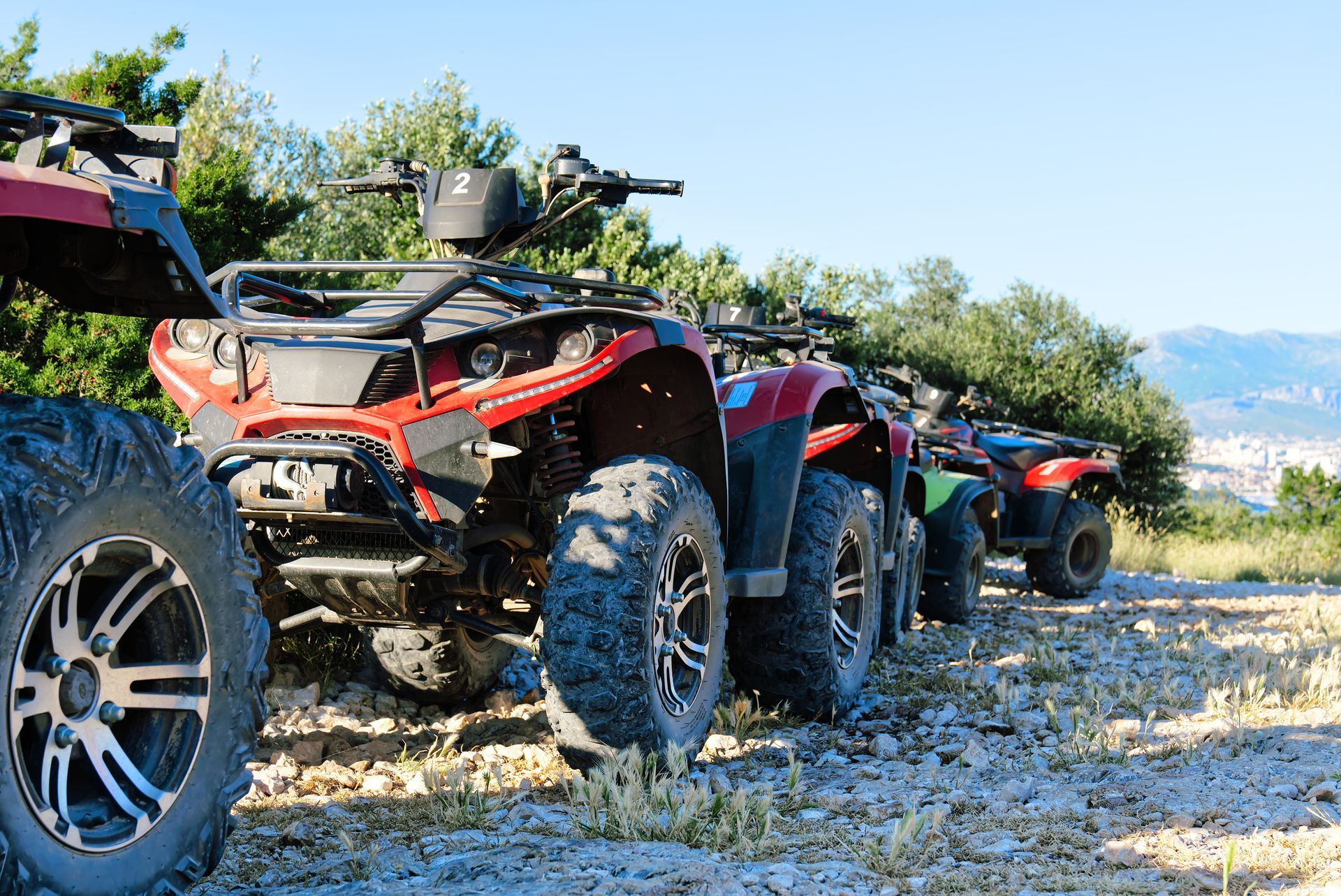 A row of red all-terrain vehicles parked on a gravel path against a clear blue sky and trees.