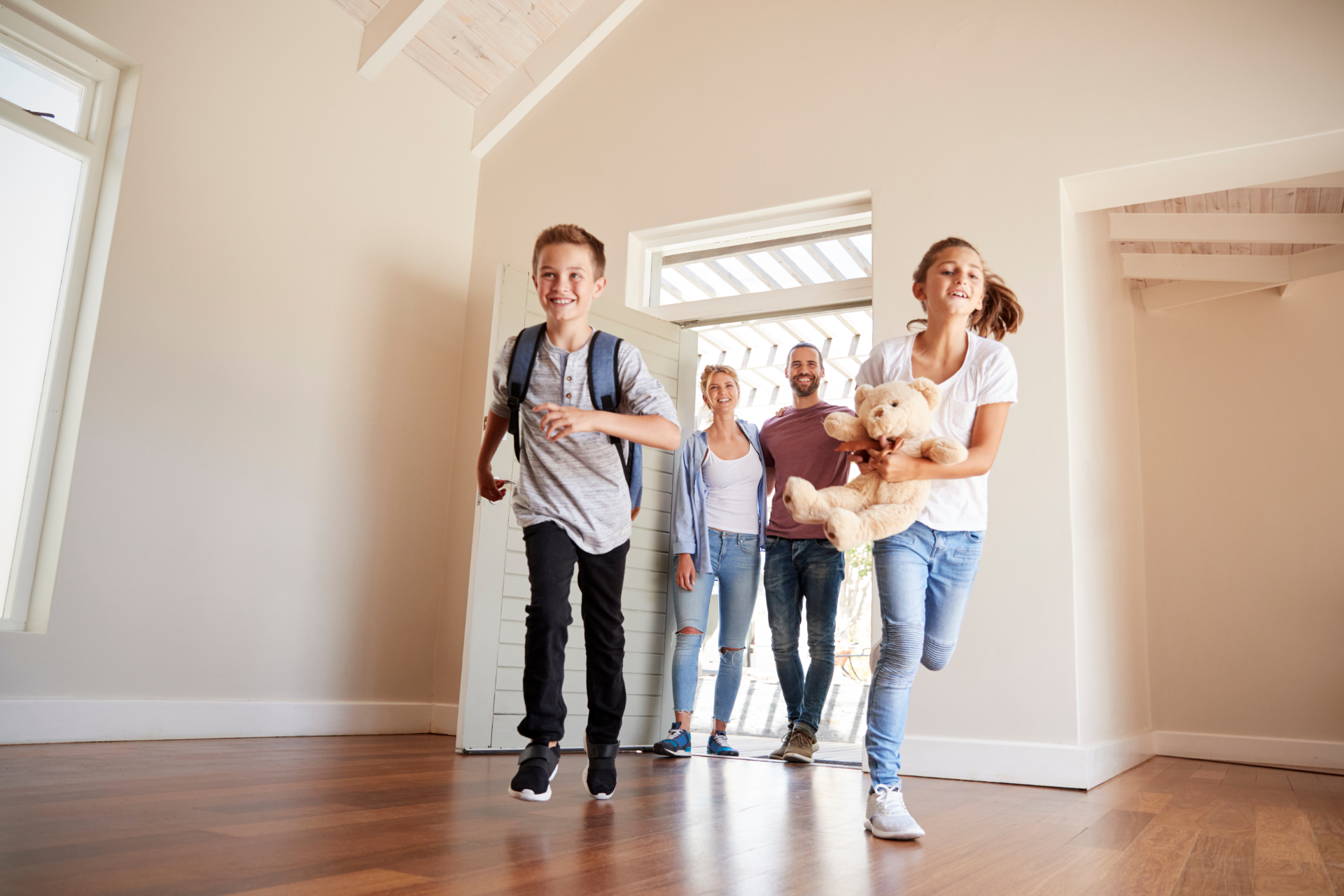A family runs excitedly into a sunlit, empty house, with two children in front and two adults following behind.