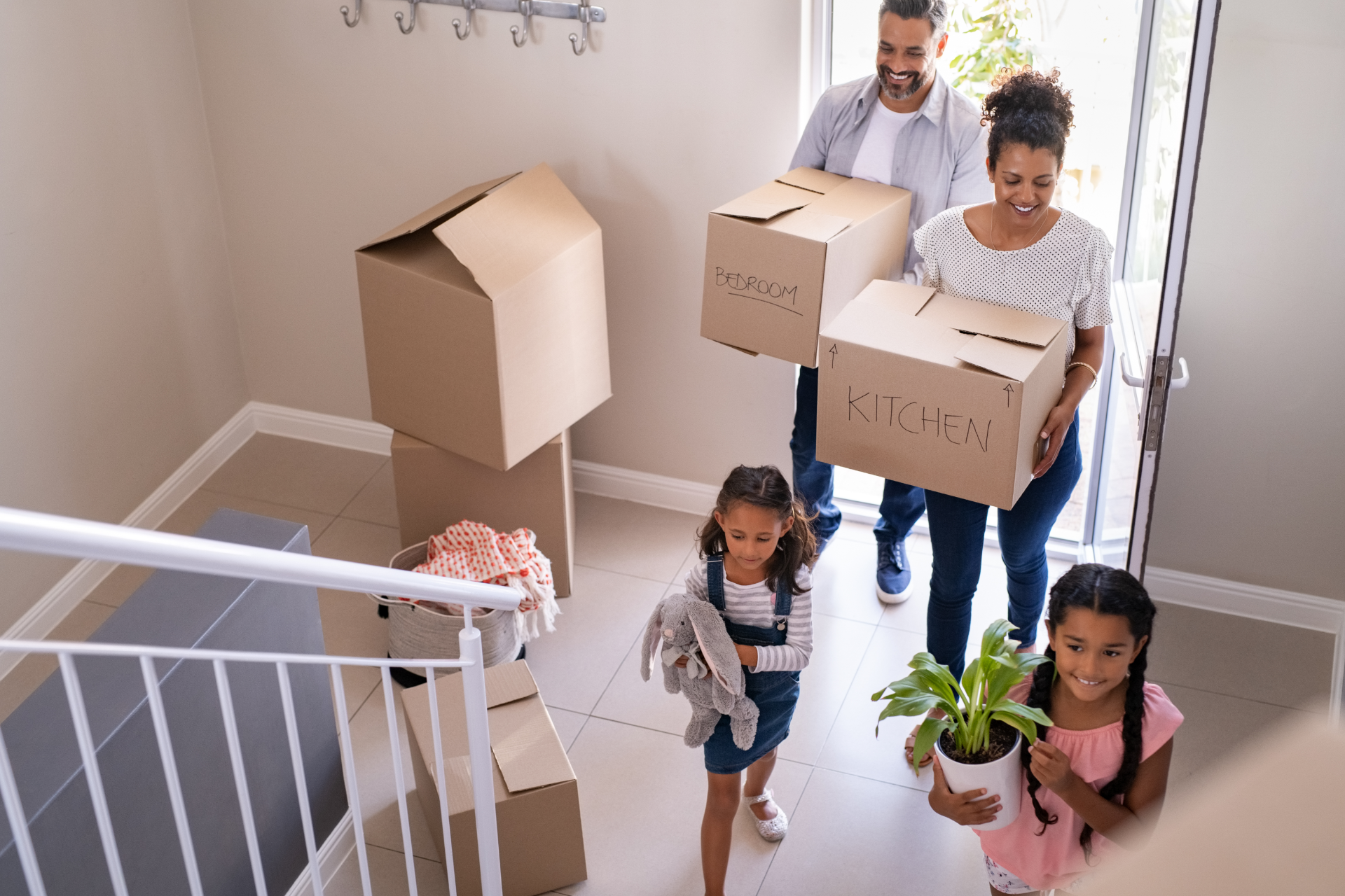 A family enters a new home carrying cardboard boxes and a houseplant, with more moving boxes in the hallway.