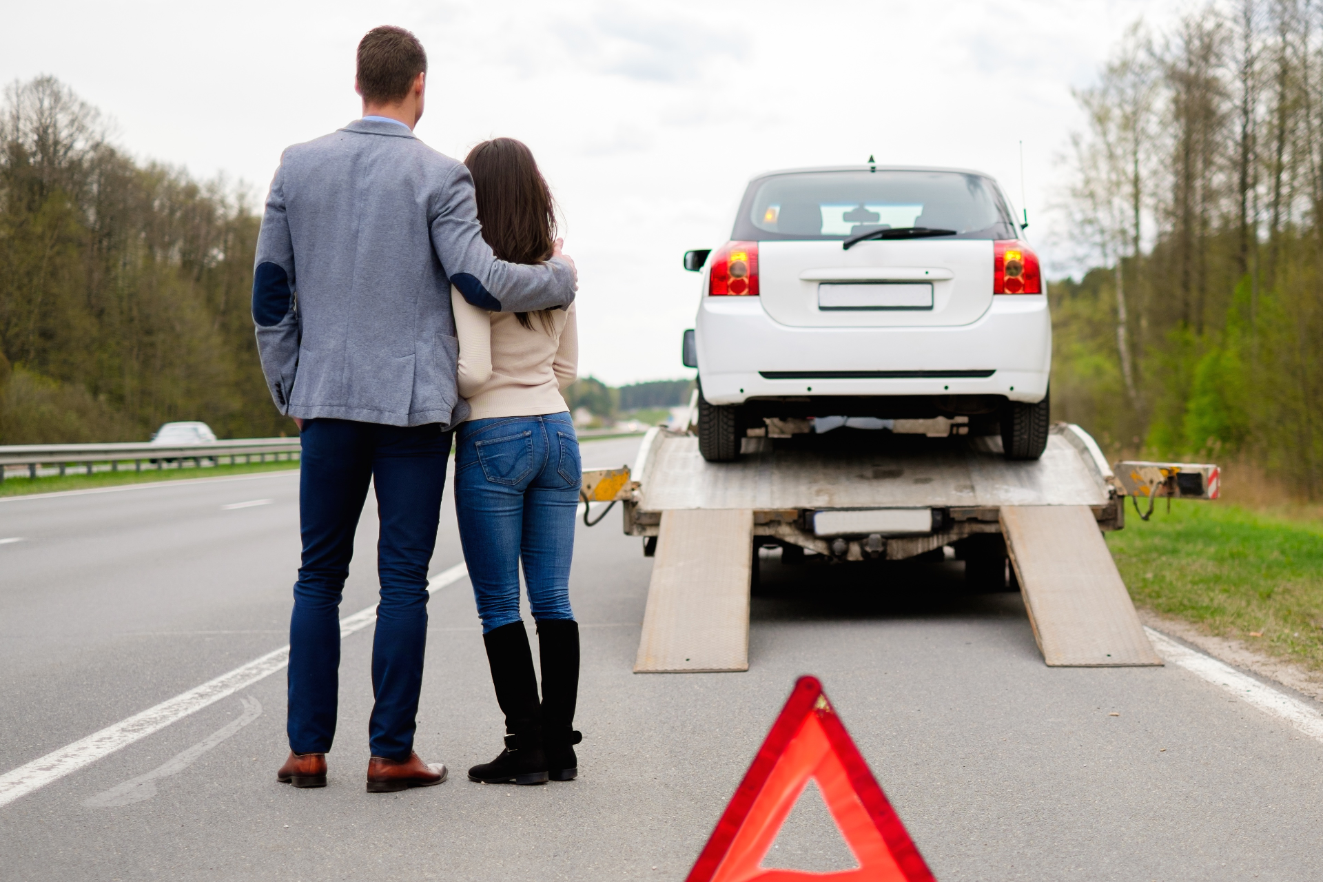 A couple stands on a highway shoulder watching their white car being loaded onto a flatbed tow truck near a warning triangle.