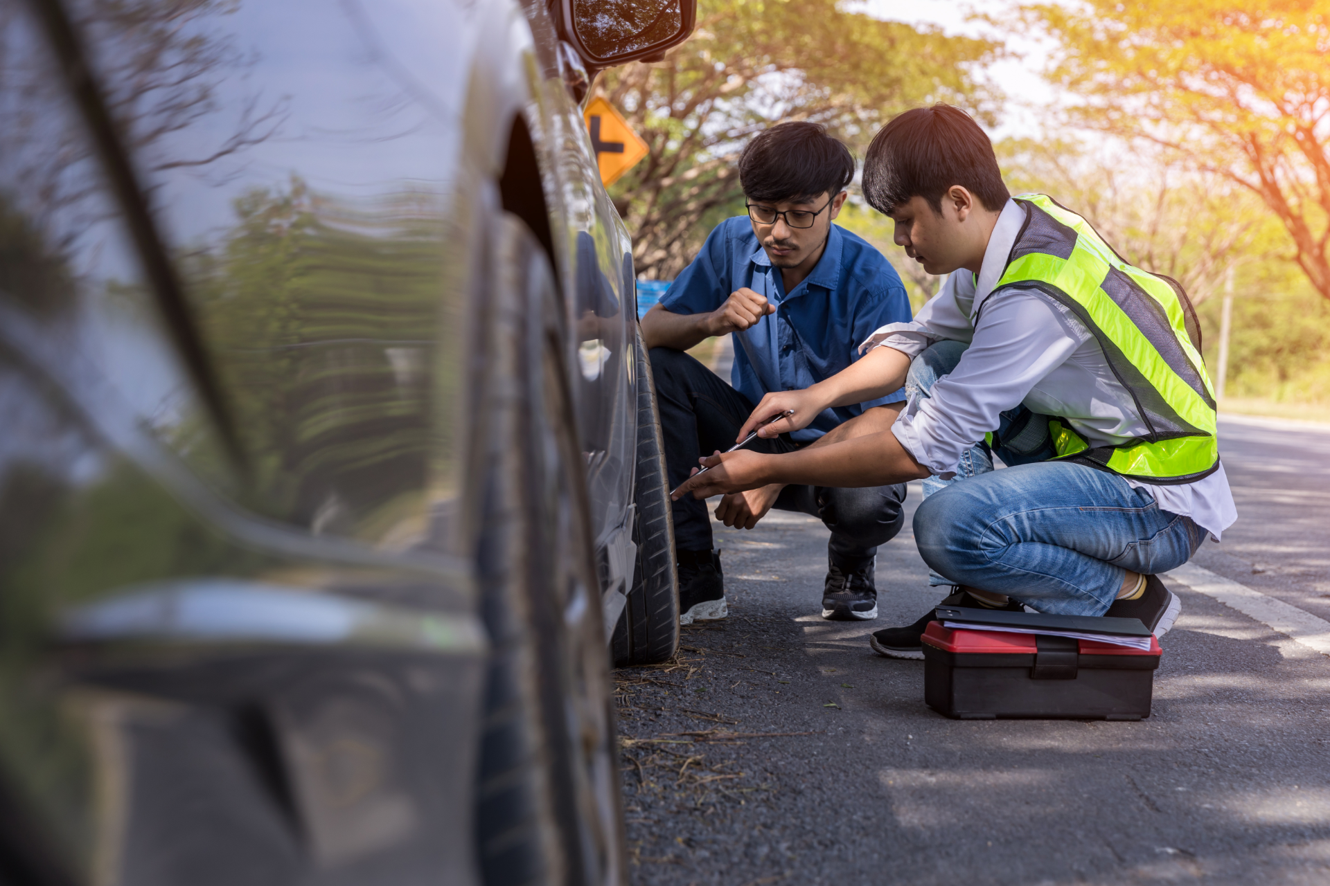 Two people in safety gear crouch by a car on the side of a road, working together to repair or change a tire.