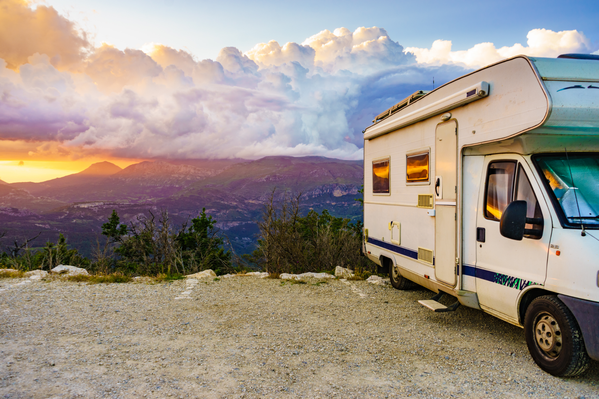A white motorhome parked on a gravel overlook facing a scenic sunset over distant mountains.
