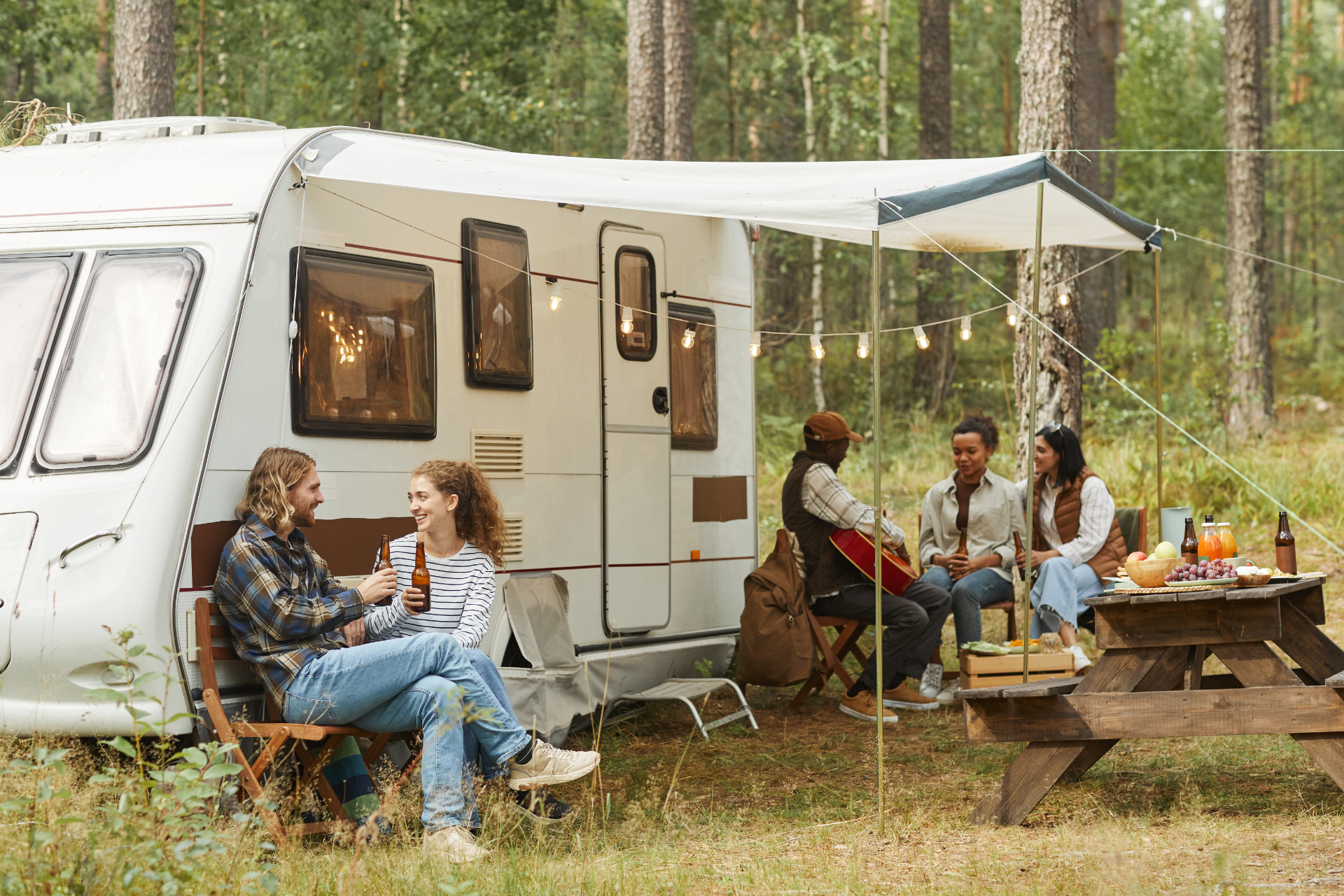 Four friends relax near a camping trailer in a pine forest, with two sitting on chairs and two at a picnic table.