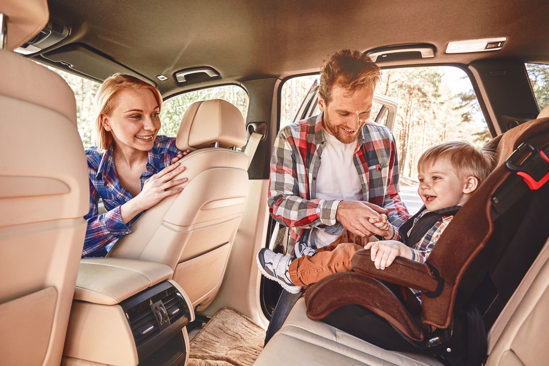 Parents buckle a smiling toddler into a car seat in the back seat of a vehicle.