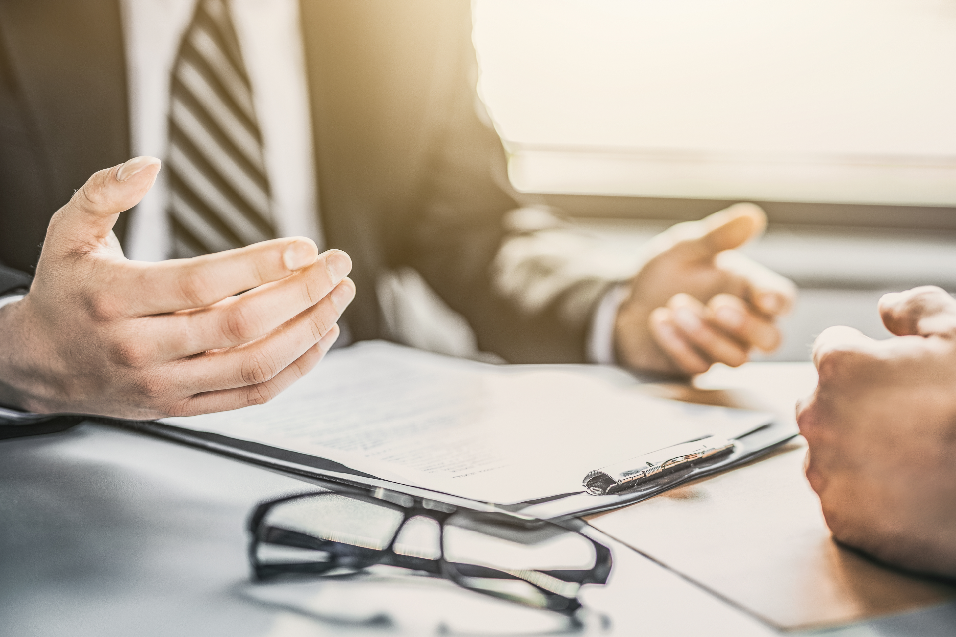 Two people in business attire gesturing during a professional meeting across a desk with documents and glasses.