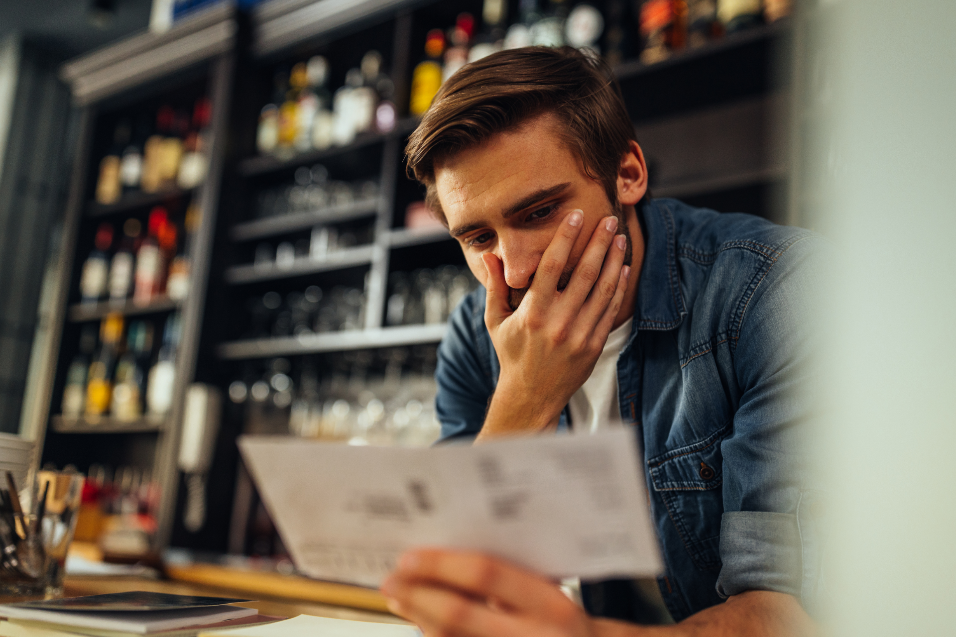 A person in a denim shirt looking at a document with a worried expression inside a bar with shelves of bottles.