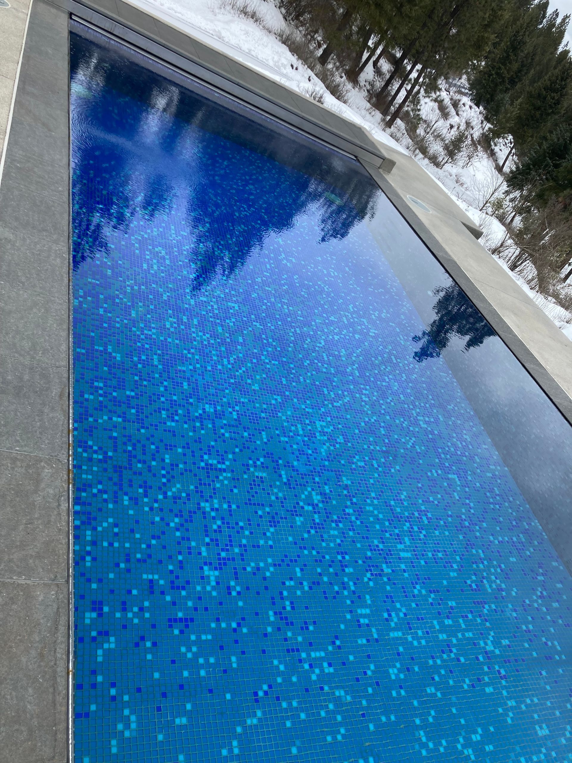 Blue tiled swimming pool reflecting trees, surrounded by stone and snow.