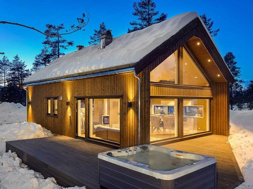 Wooden cabin at dusk with snow-covered roof, large windows, and lit jacuzzi; trees in background.