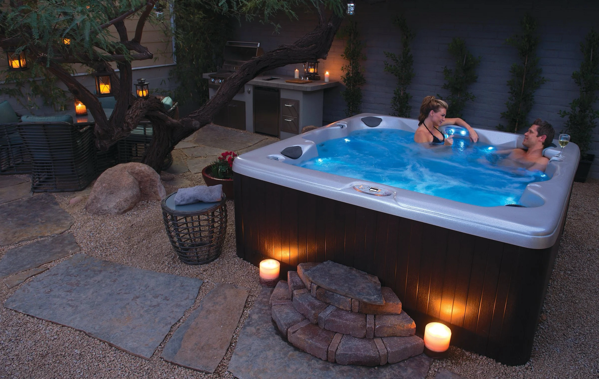 Two women relaxing in a lit-up hot tub outdoors, surrounded by candles and a small zen garden.