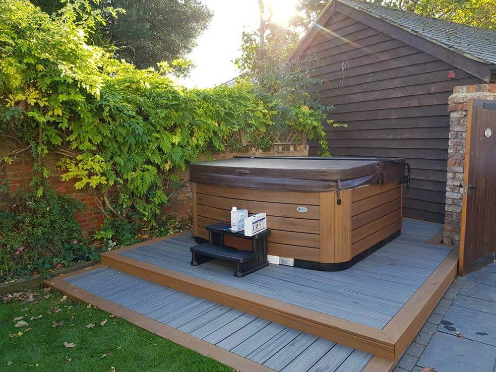 Hot tub on a wooden deck next to a building and lush greenery. Brown and gray tones, sunny outdoors.