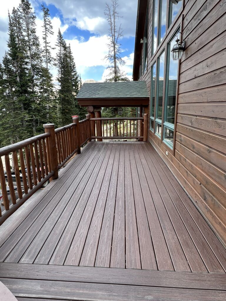 Wooden deck with railing overlooking trees, connected to a cabin with large windows.