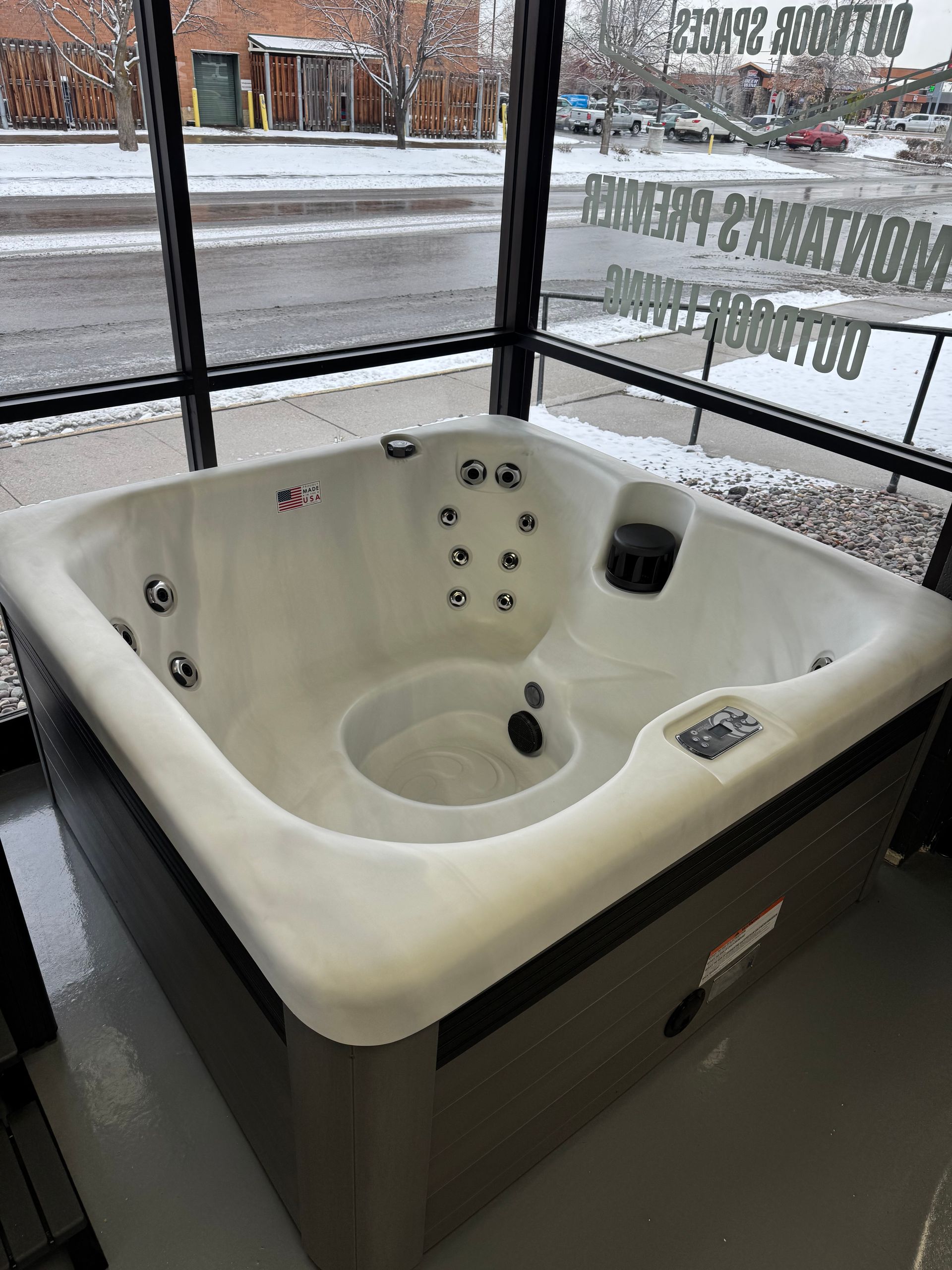 A square, white hot tub with multiple jets, inside a showroom, with a window and snowy outside view.