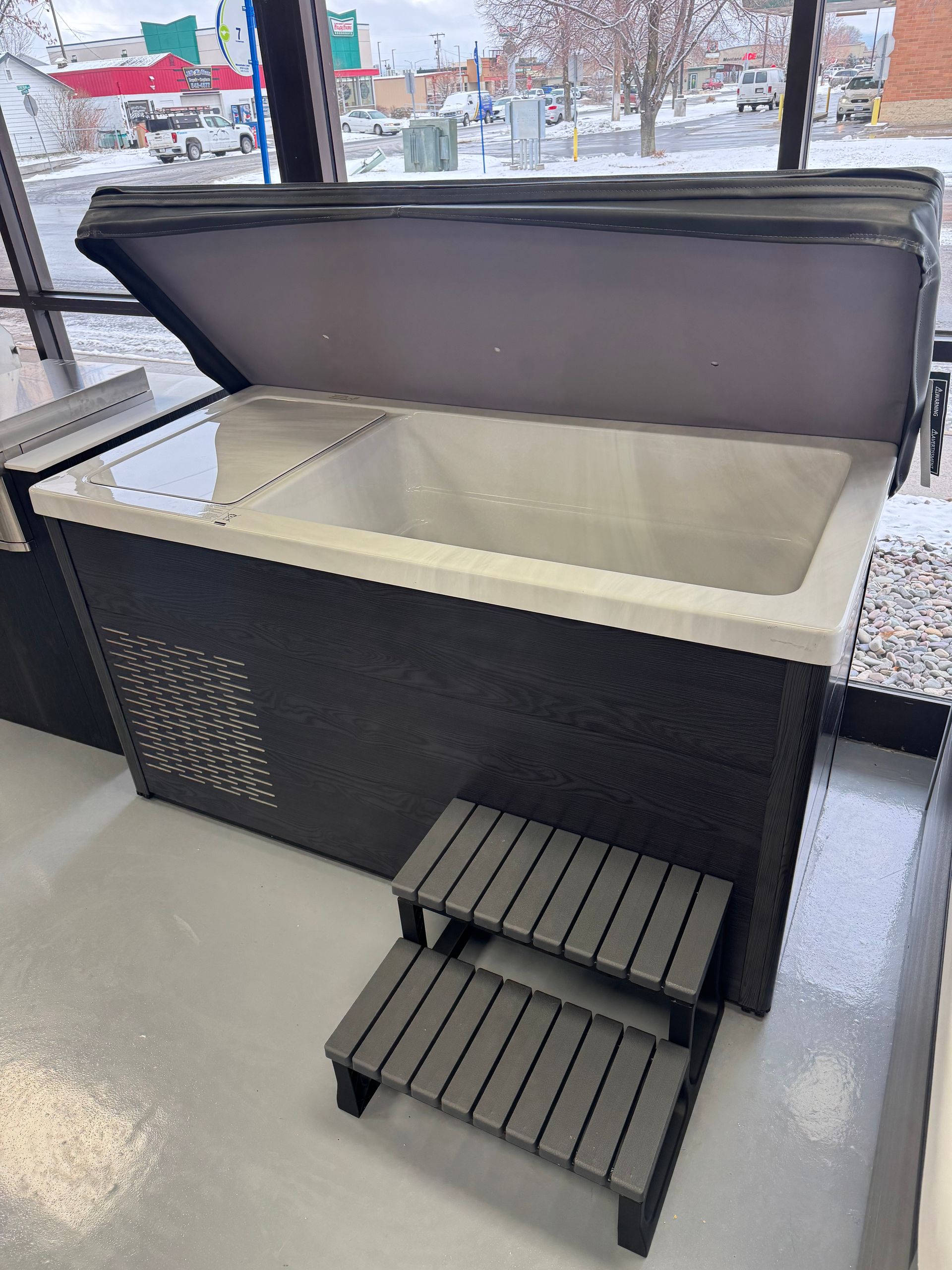 Hot tub with open cover, steps in front. Black and gray exterior, white interior.