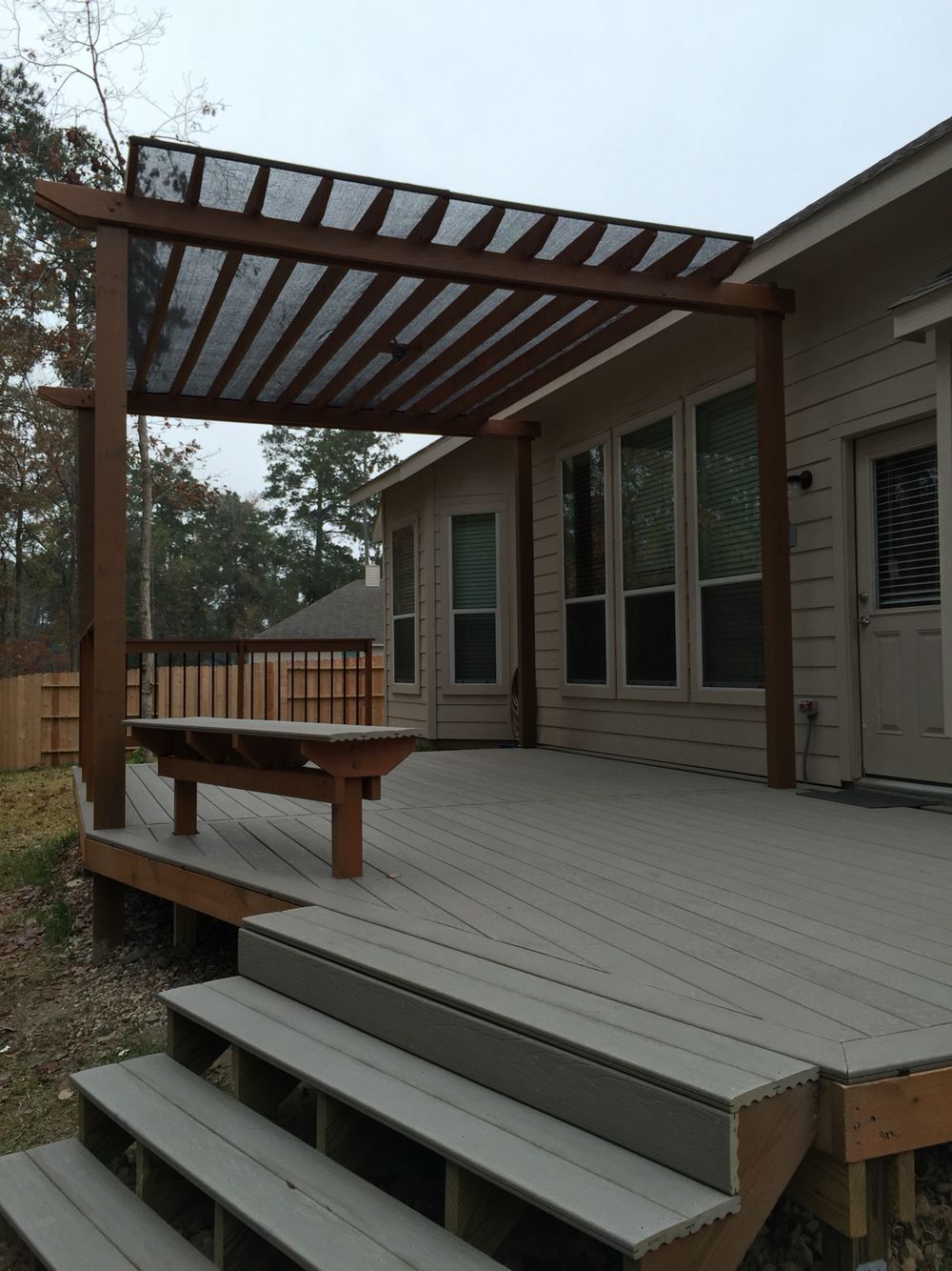Wooden deck with steps, pergola with shade, and attached to a light-colored house. Bench under the pergola.