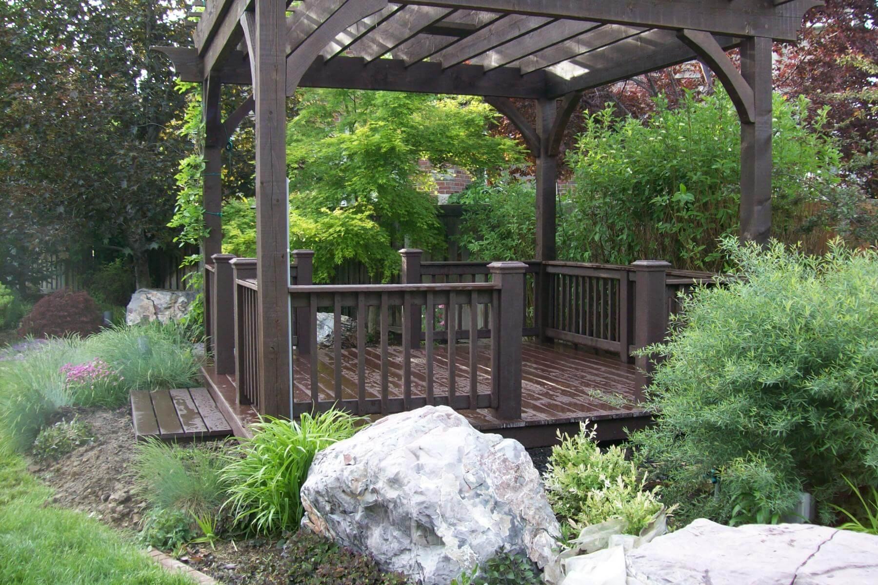 Wooden gazebo in a garden surrounded by green bushes and rocks.