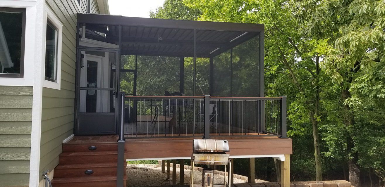 A screened-in porch with a dark railing and roof attached to a light green house, surrounded by trees.