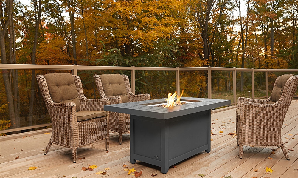 Outdoor deck with three wicker chairs around a lit fire pit. Autumn foliage in background.