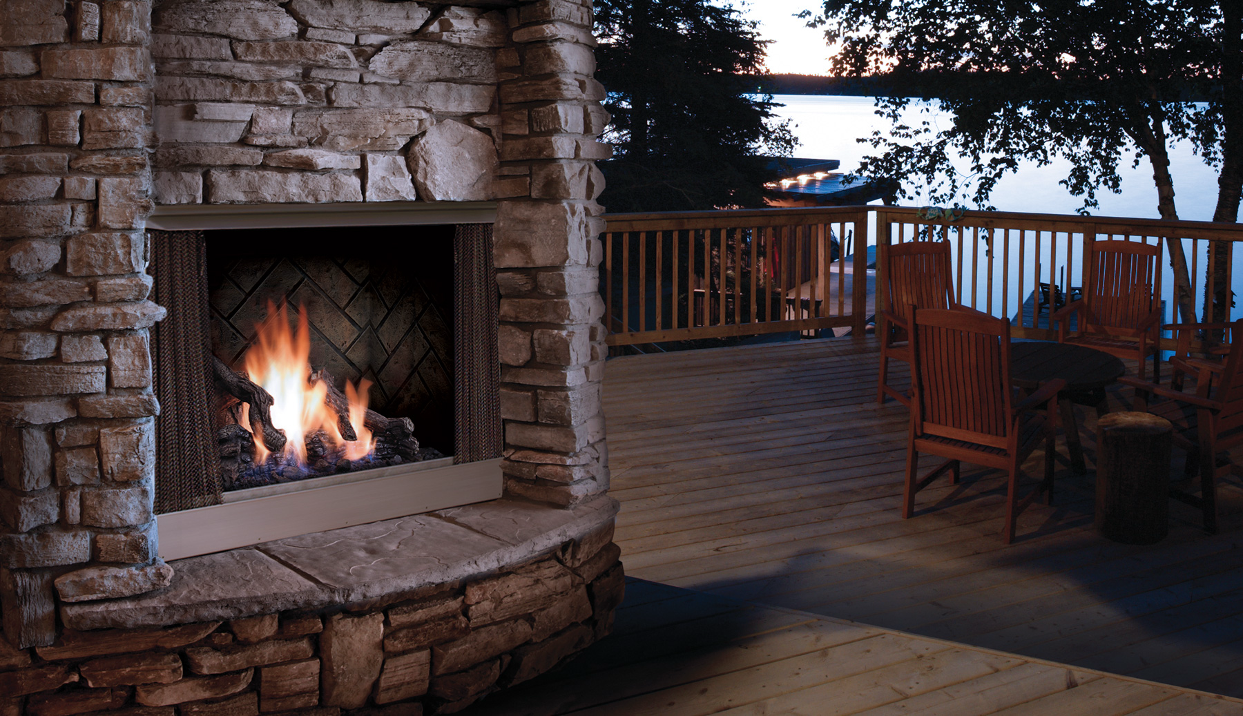 Outdoor stone fireplace with flames, next to wooden deck with lake view.