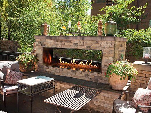 Outdoor stone fireplace with flames, seating area, and decorative plants.