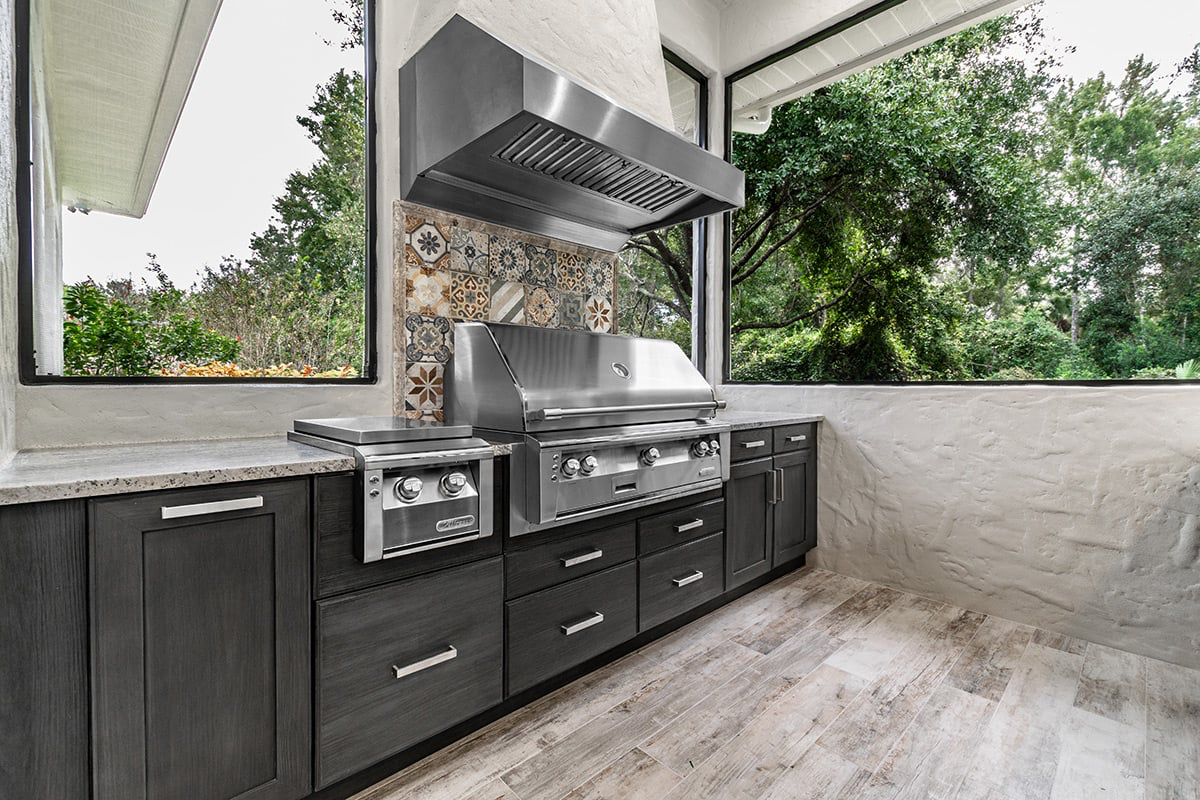 Outdoor kitchen with stainless steel grill, cabinets, and windows overlooking greenery.