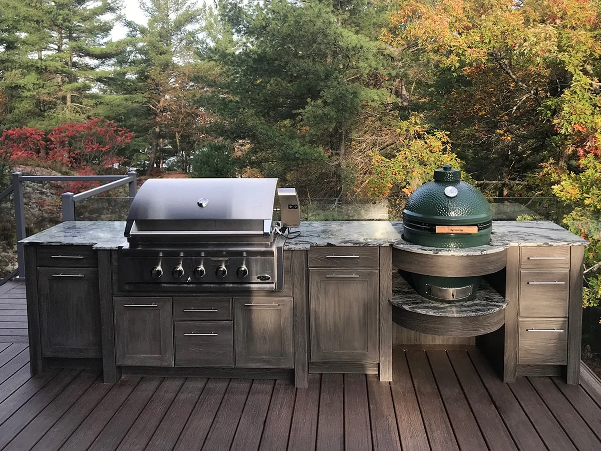 Outdoor kitchen with a gas grill and a Big Green Egg, set on a wooden deck with a forest background.