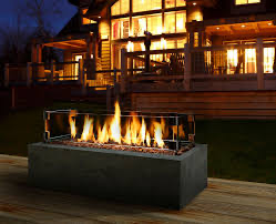 A rectangular gas fire pit with flames, in front of a lit wooden house at night.