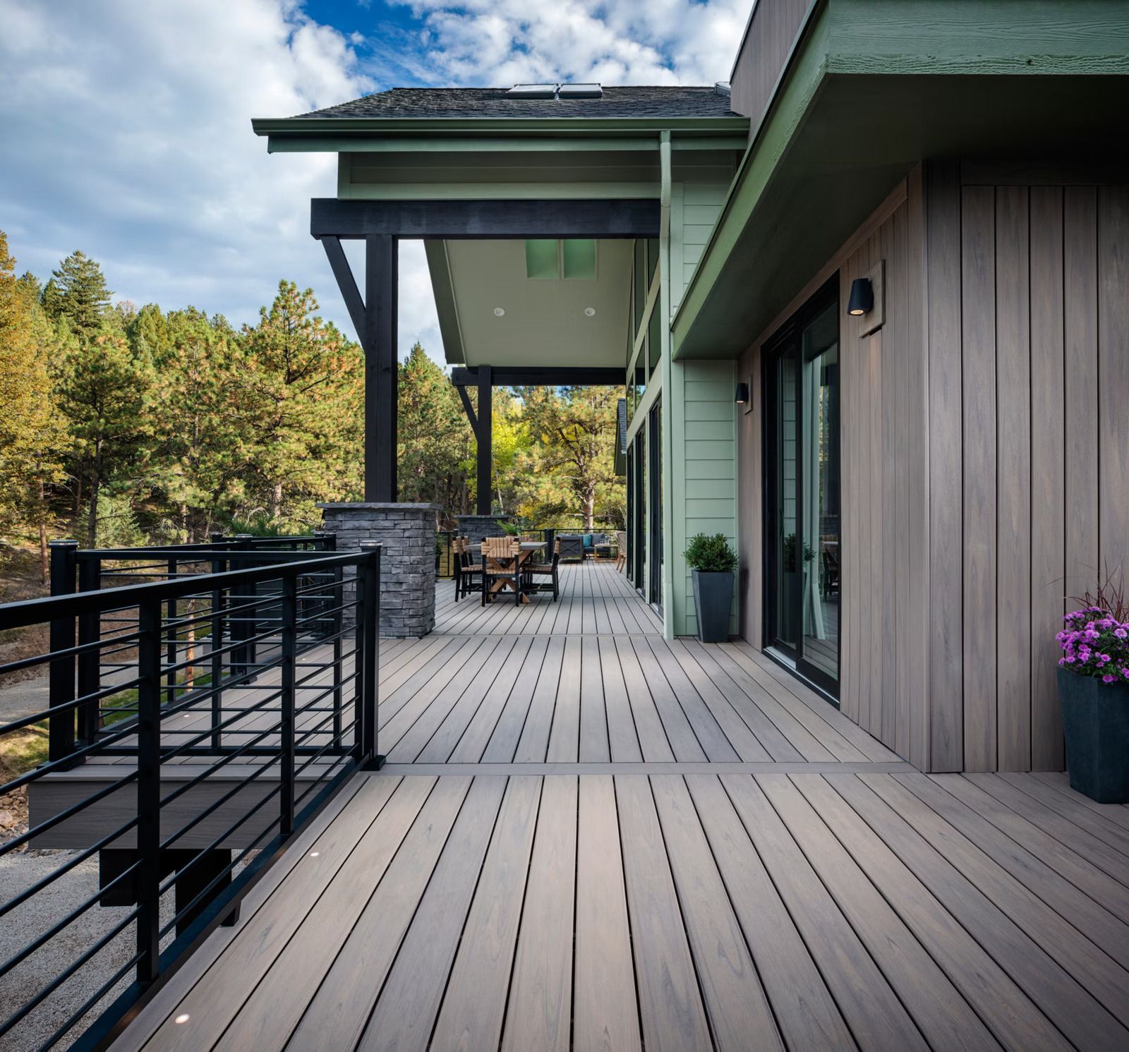 Wooden deck with black railing, overlooking a forest. Green house with patio furniture.