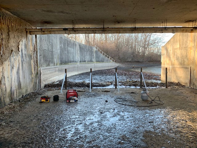 A muddy area under a bridge with a generator and tools on the ground.