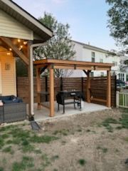 A wooden outdoor pergola covers a black grill station and workspace on a concrete patio next to a house.