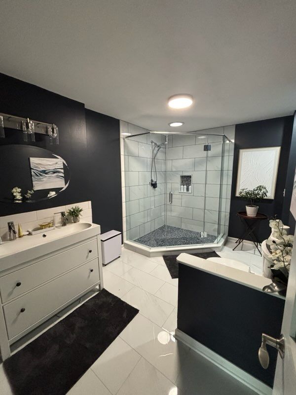 Dark blue bathroom with white vanity, large glass shower, black rug, and white floor tiles.
