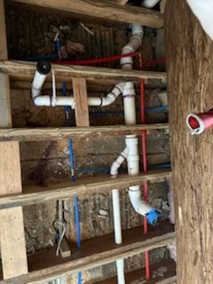 White PVC drainage pipes and blue and red PEX water lines installed between wooden floor joists in an open wall cavity.