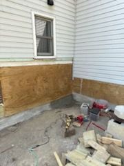 A home construction site with plywood walls attached to a siding exterior and loose wood scraps scattered on the ground.