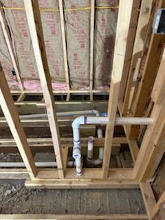 White PVC plumbing pipes installed within an unfinished wooden wall frame in front of fiberglass insulation.