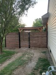 A horizontal wooden slat fence with black metal posts extends from the side of a house toward a large tree in a yard.