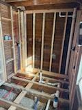 An interior view of a room under construction, showing wooden wall studs, floor joists, and exposed white plumbing pipes.