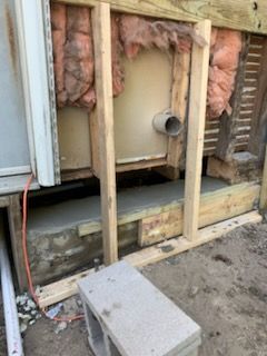 A wooden wall frame sits over a concrete foundation next to an exposed wall with pink insulation and a protruding pipe.