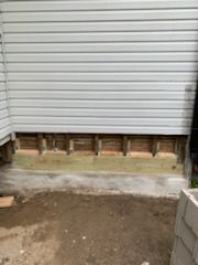 A view of a house exterior showing light gray siding above an exposed wooden frame and a new green pressure-treated beam.