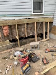 Construction framing work underway beneath a house exterior with scattered power tools and building materials nearby.
