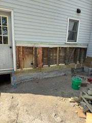 An exterior view of a house wall with siding removed to expose wooden framing, insulation, and the home's foundation.