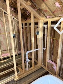 Unfinished wall framing with exposed pink insulation and white PVC plumbing pipes in a residential construction space.