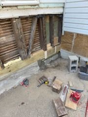 Construction materials and tools lie on the ground in front of an exterior wall with exposed wooden framing and siding.