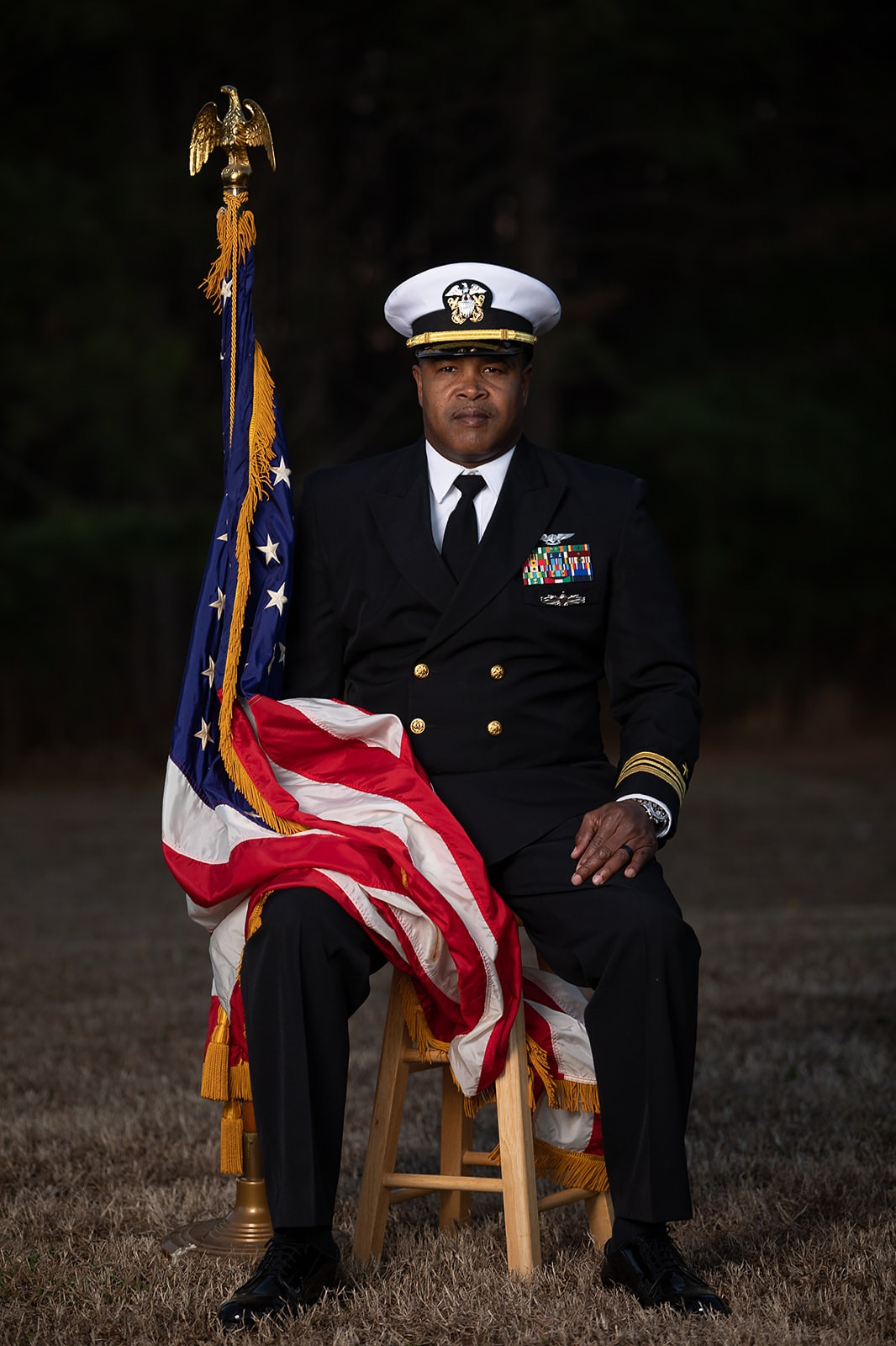 Navy officer in dress uniform seated, holding American flag. Outdoors, dark background.