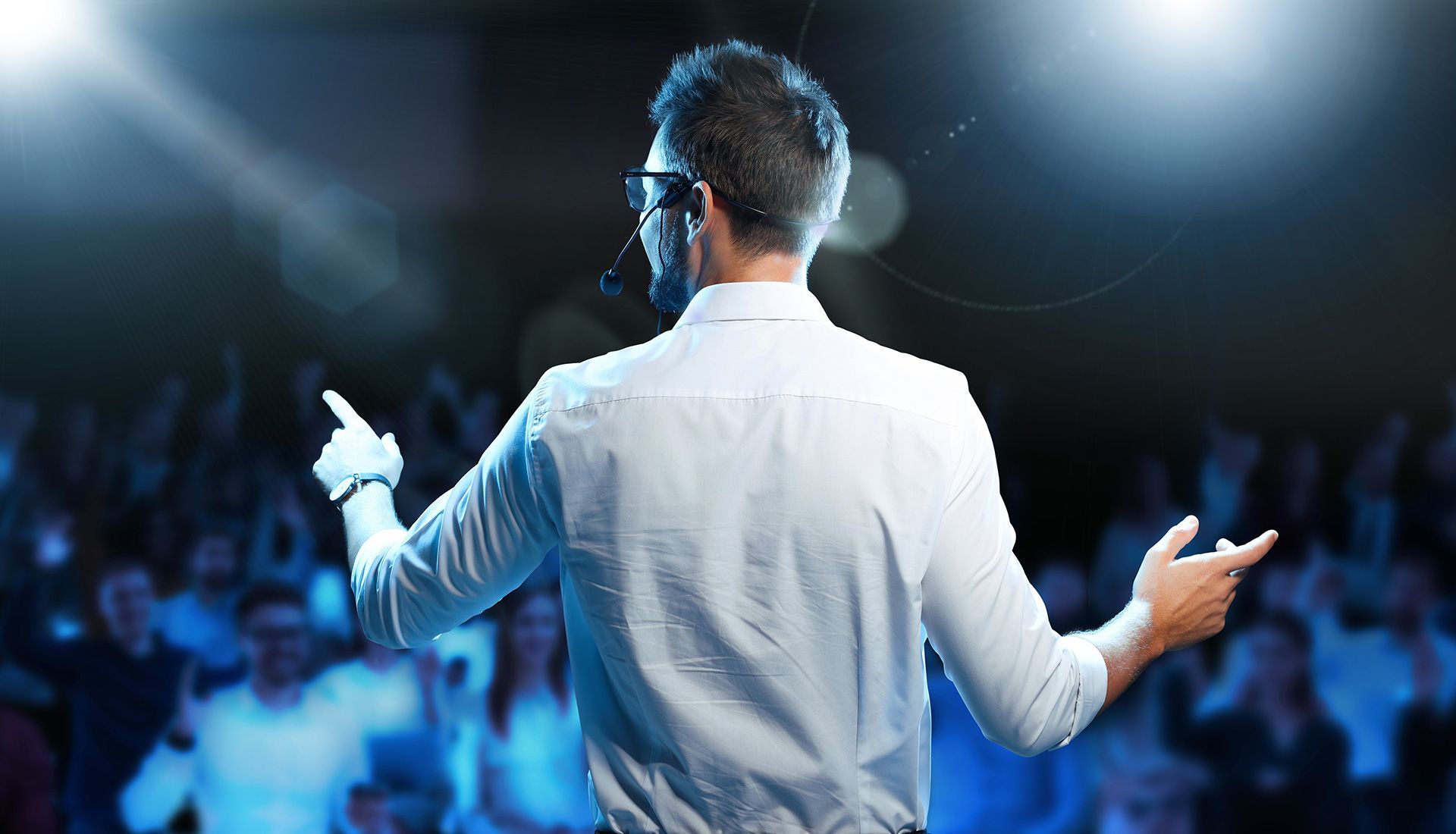 Man on stage with microphone, gesturing to a crowd. He is wearing a white shirt; stage is lit with blue light.